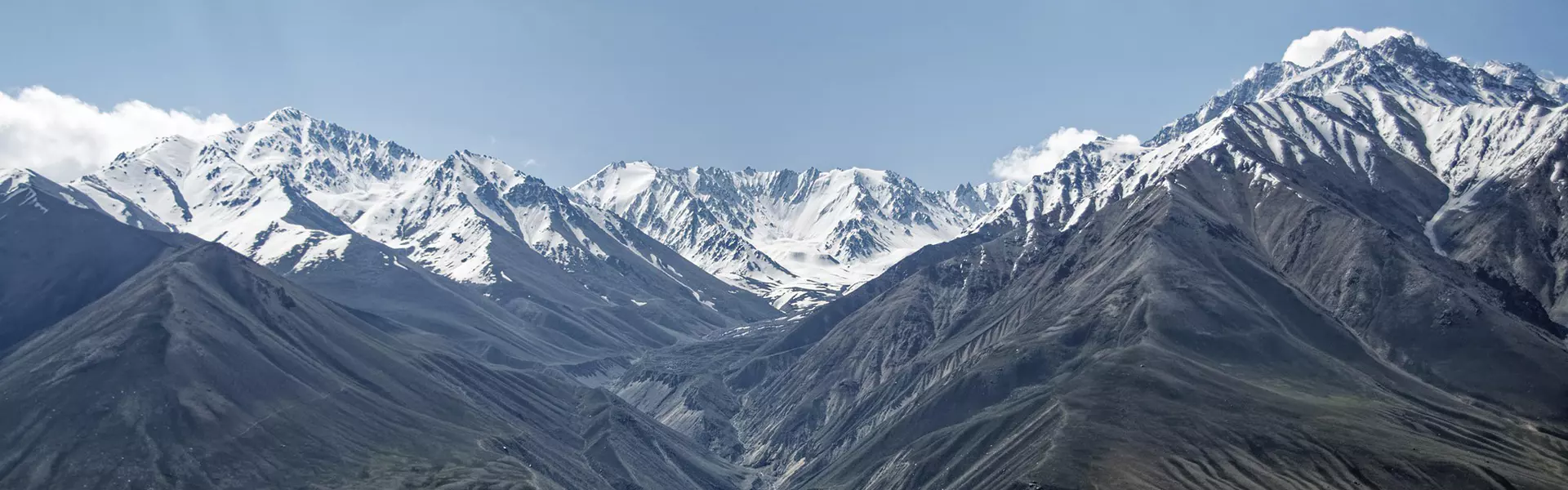 Onder een blauwe hemel met hier en daar wat wolken rijzen besneeuwde bergtoppen op, terwijl beneden een kronkelende rivier door de ruige, rotsachtige vallei stroomt.
